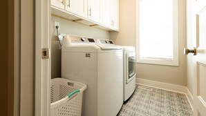 A laundry room with two front-loading washing machines, a white laundry basket, and a window with blinds. - Three Seas at the B, Walk to Tiki Bar and Beach (Carolina Beach)