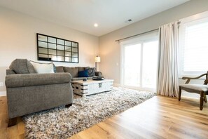 A living room with a grey sofa, a wooden coffee table, a fluffy rug, and a large window with curtains.