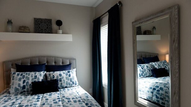 A bedroom with a bed, a headboard, a mirror, and a window with curtains. - Three Seas at the B, Walk to Tiki Bar and Beach (Carolina Beach)