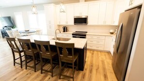 A modern kitchen with white cabinetry, a stainless steel refrigerator, and a dining area with wooden chairs. - Three Seas at the B, Walk to Tiki Bar and Beach (Carolina Beach)
