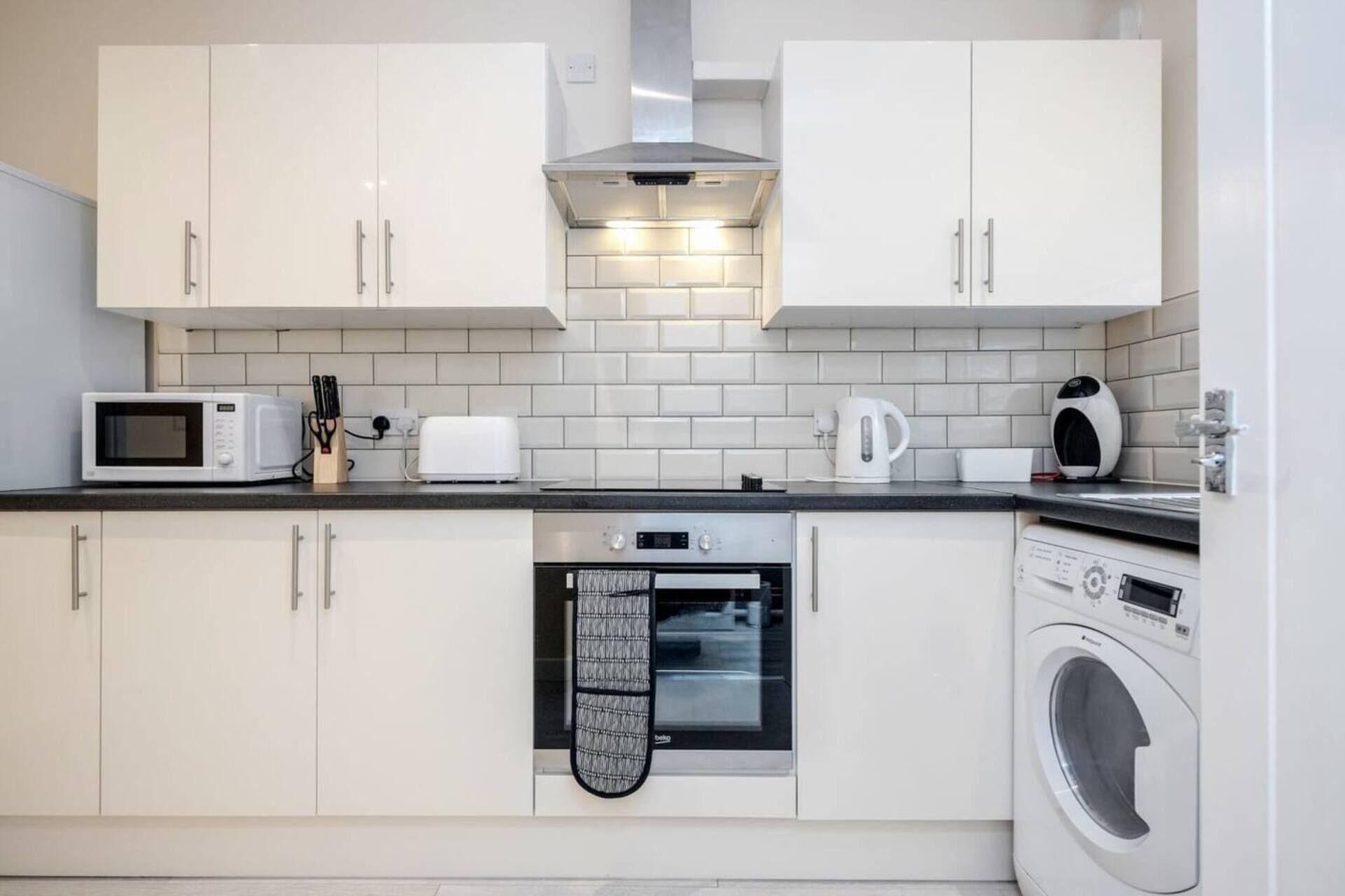 A compact kitchen with white cabinets, a microwave, toaster, kettle, oven, and a washing machine.