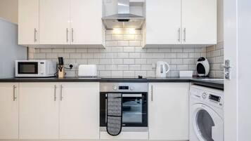A compact kitchen with white cabinets, a microwave, toaster, kettle, oven, and a washing machine.