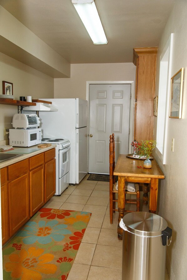 A compact kitchen with a microwave, stove, and refrigerator. There's a wooden table with a trash can and a chair. A colorful rug is on the floor. - Langridge Lodge Studio Residence by RedAwning (Alpine)