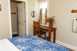 Un dormitorio con una cama, un escritorio de madera, una silla, un ventanal con cortinas, un espejo y un perchero en la pared.