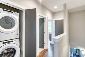 Een wasserette met gestapelde wasmachines en drogers, een gang naar een slaapkamer en een uitzicht op een woonruimte met een plant.