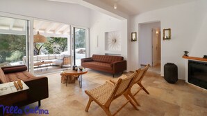 Ein geräumiger Wohnbereich mit braunem Sofa, Korbstühlen und einem Couchtisch. Ein Kamin und ein großes Fenster sind zu sehen.