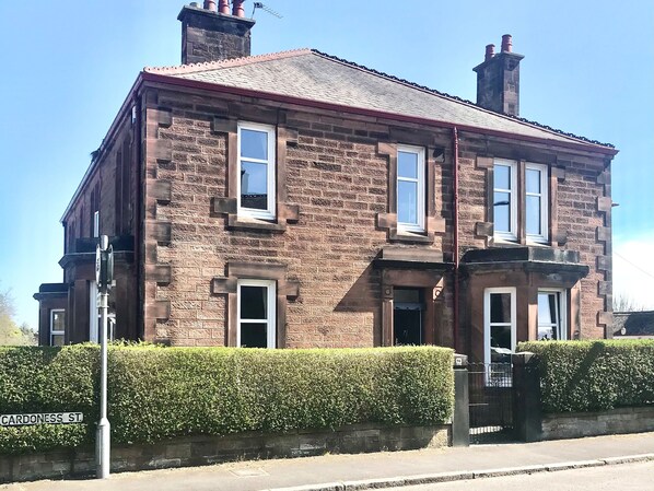 A two-story stone building with multiple windows, a brick chimney, and a hedge in front. - 4 bedroom accommodation in Dumfries (Dumfries)