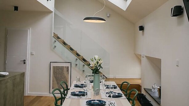 Un comedor moderno con techo alto, tragaluces y una mesa de madera preparada para cuatro comensales.