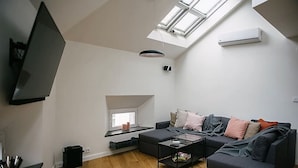 Una sala de estar moderna con un sofá grande de color gris, una mesa de centro y un televisor montado en la pared. Hay un tragaluz sobre el sofá y un equipo de aire acondicionado en la pared.