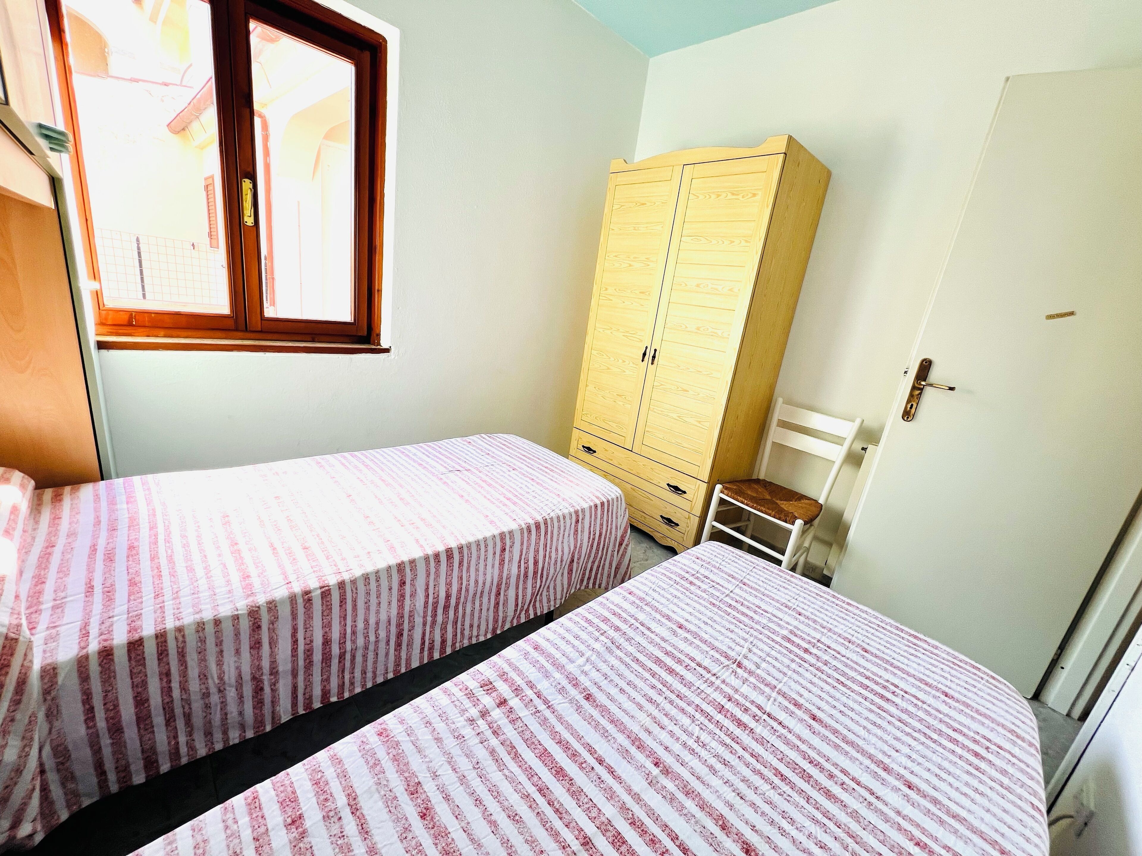 A bedroom with two beds, a wooden wardrobe, a chair, and a window.