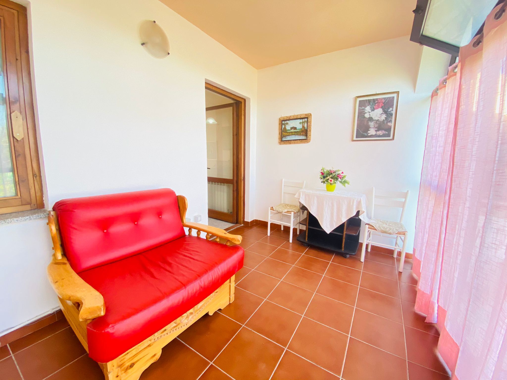 Un canapé rouge, une petite table avec un vase et une chaise dans une pièce dont le sol est recouvert de carreaux et qui possède une porte.