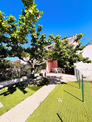 A courtyard with a paved pathway, a small table and chairs, and a tree providing shade.