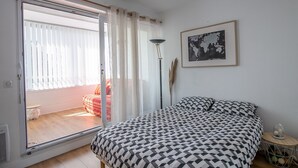 A bedroom with a bed, a window with curtains, a floor lamp, a framed map, and a small side table with decorative items.