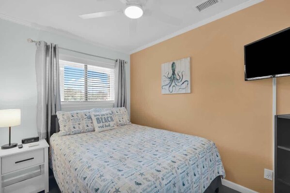 A bedroom with a bed, a nightstand, a lamp, a window with blinds, and a wall-mounted TV.