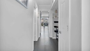 Corredor moderno e bem iluminado, com paredes brancas e piso ladrilhado, que dá acesso a um quarto com cama e escrivaninha.