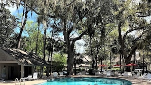 A swimming pool with lounge chairs and umbrellas, surrounded by trees and a building.