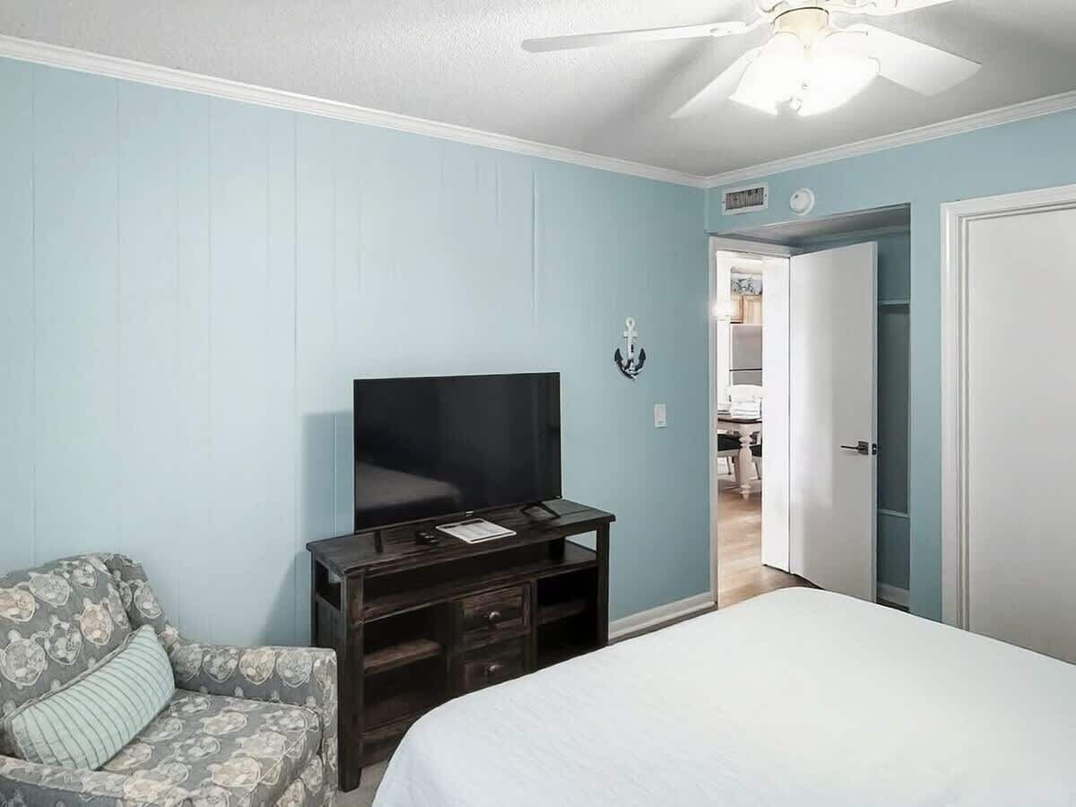 Un dormitorio con una cama, un televisor sobre un mueble, una silla y una puerta que da a otra habitación.