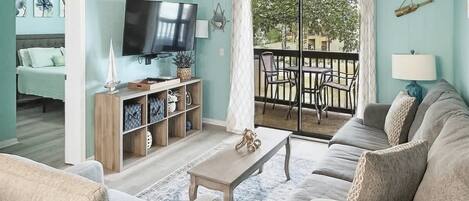 Sala de estar con sofá, mesa de centro, televisor y estantería. Hay un balcón con mesa y sillas.