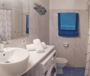 Un bagno con un lavandino bianco, uno specchio, un portasciugamani con asciugamani, un water e un asciugamano blu appeso al muro.