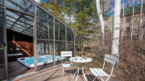 Un espace de détente avec un jacuzzi entouré de parois de verre, doté d’une terrasse en bois, de chaises blanches et d’un chalet en bois en arrière-plan.