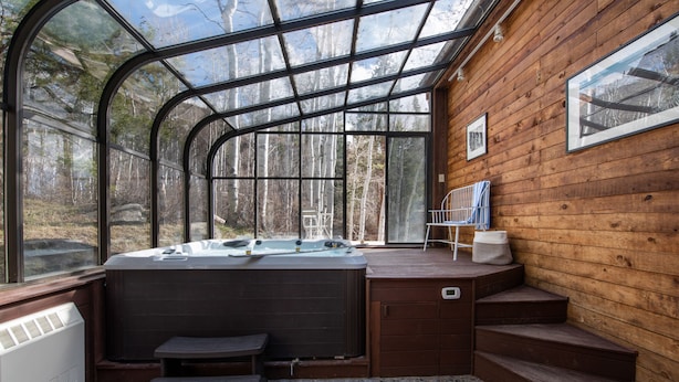 Un bain à remous doté d’une terrasse en bois et d’escaliers, entouré d’une enceinte en verre offrant une vue sur la forêt.
