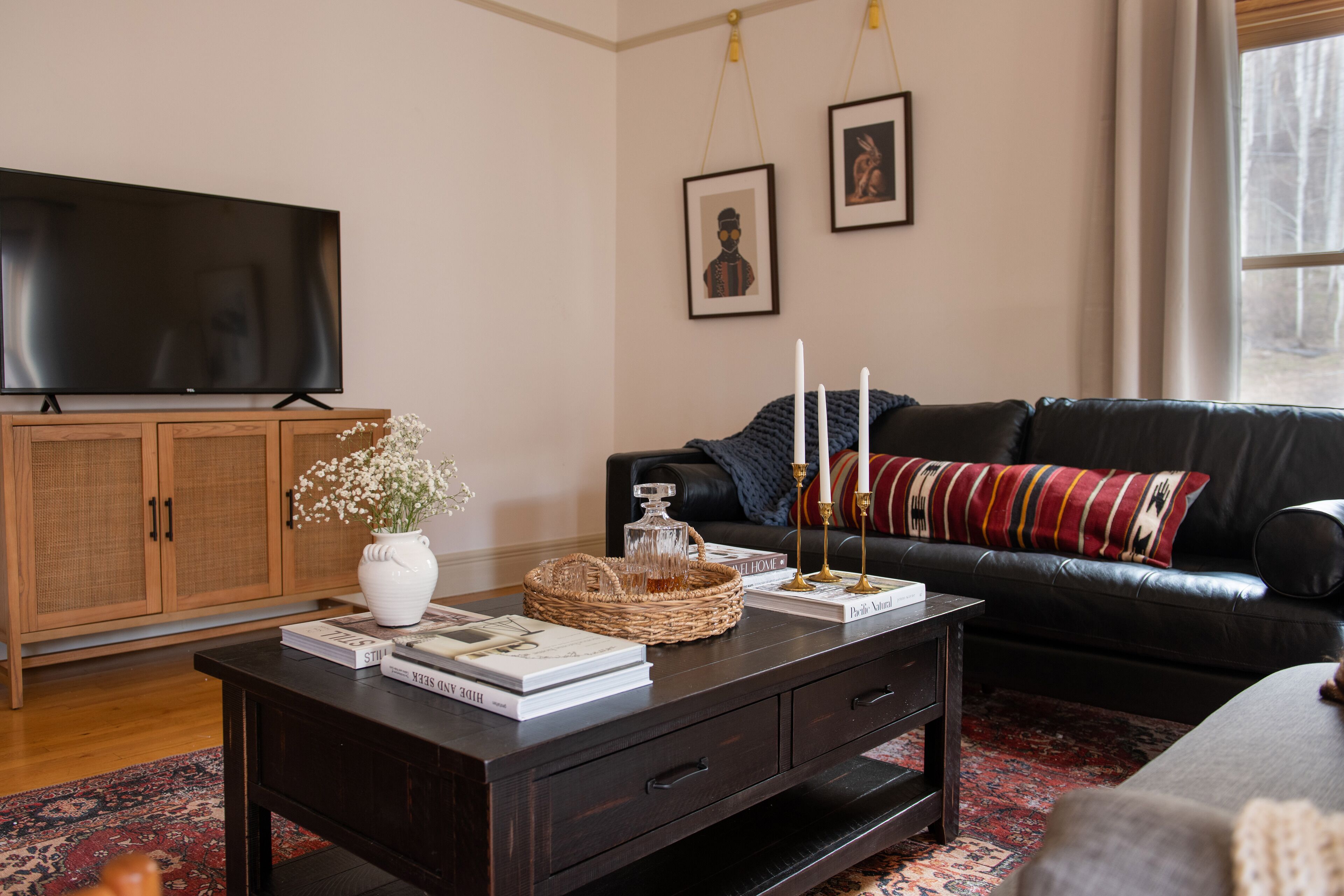 Un salón con una mesa de centro de madera oscura, un sofá de cuero negro, un televisor sobre un mueble de madera y un ventanal con vistas a árboles.
