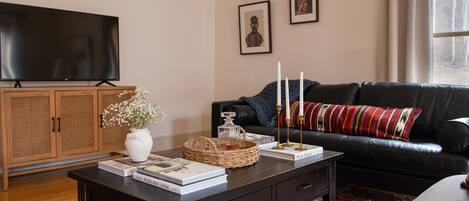Un salón con una mesa de centro de madera oscura, un sofá de cuero negro, un televisor sobre un mueble de madera y un ventanal con vistas a árboles.