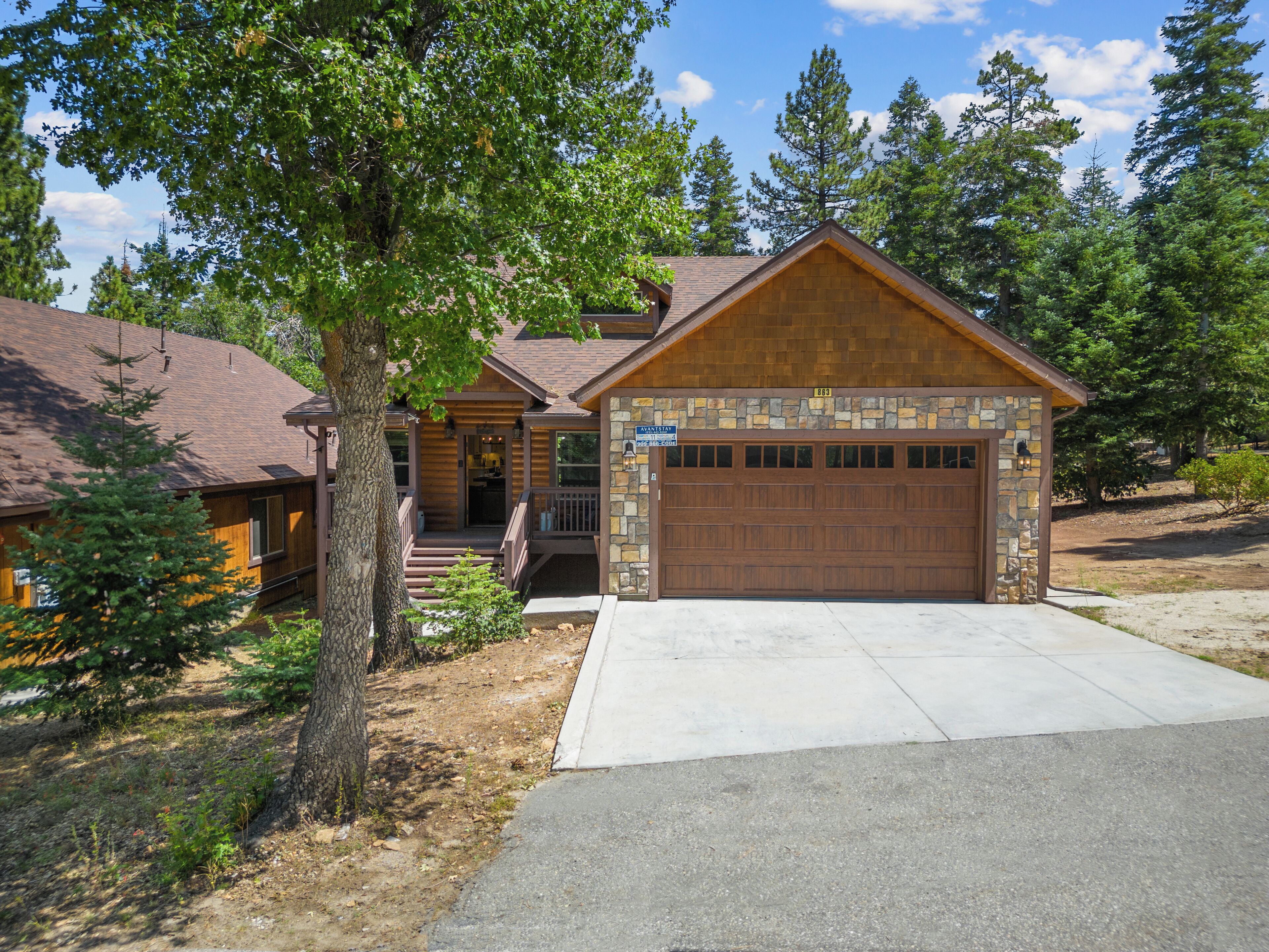 A two-story log cabin with a stone facade garage, surrounded by trees and a clear sky.