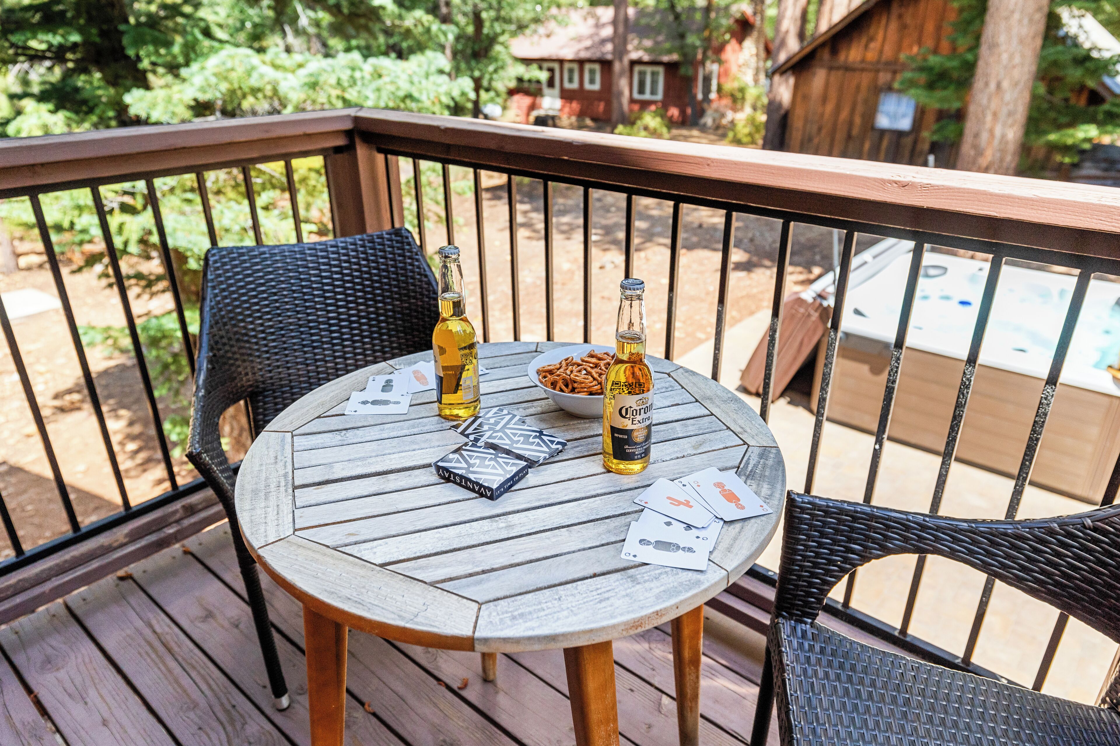 A round wooden table with two bottles, a bowl of snacks, and playing cards on a decked outdoor area with wicker chairs.