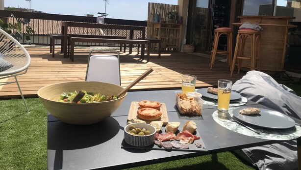 Un área de comedor al aire libre con una mesa preparada para el almuerzo, que incluye ensalada, pan, queso, aceitunas y bebidas.