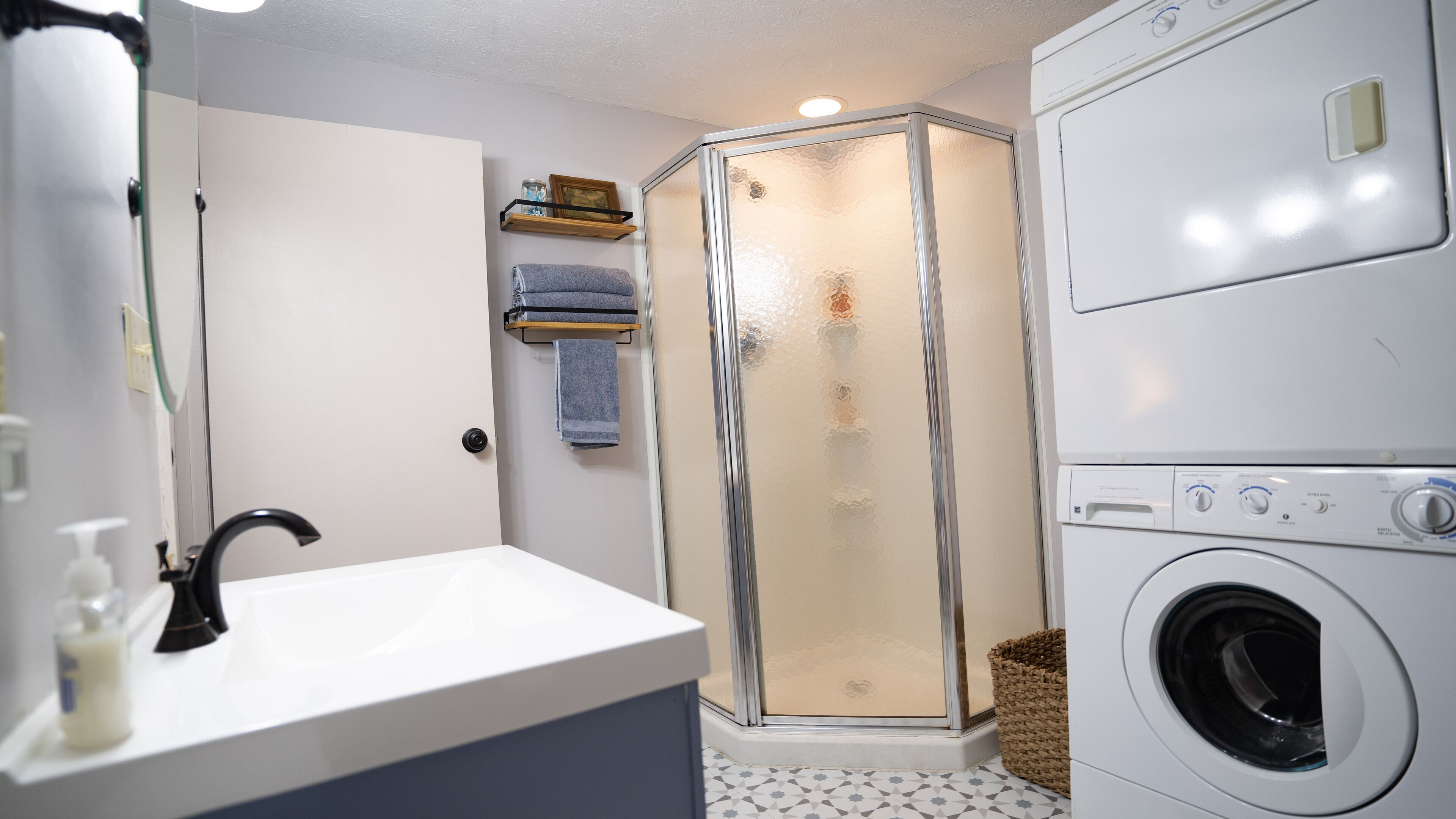 A compact bathroom with a sink, mirror, shower enclosure, and a front-loading washing machine.