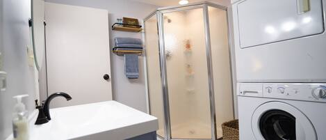 A compact bathroom with a sink, mirror, shower enclosure, and a front-loading washing machine.