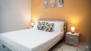A neatly made bed with white linens and two patterned pillows, a bedside table with a lamp, and three framed pictures on the wall. - Malta's Beachfront Apartment in Mellieha (Il-Mellieha)