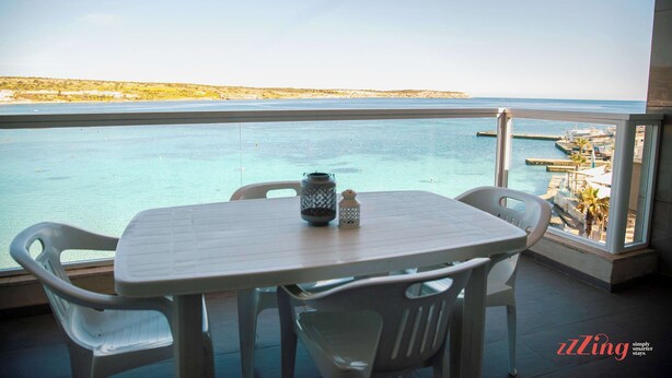 A balcony with a table and chairs overlooking a body of water and a distant shoreline. - Malta's Beachfront Apartment in Mellieha (Il-Mellieha)
