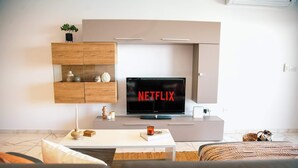 A modern living room with a flat-screen TV displaying the Netflix logo, a wooden cabinet with decorative items, a white coffee table with a candle and books, and a grey sofa. - Malta's Beachfront Apartment in Mellieha (Il-Mellieha)
