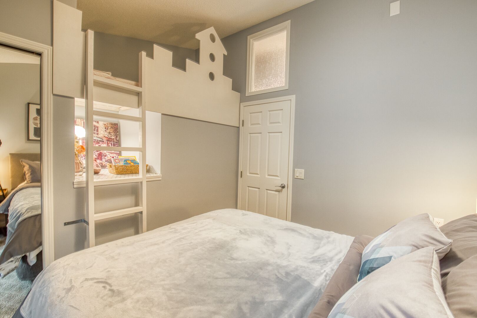 A bedroom with a bed, a ladder, a door, and a shelf with books and a basket.