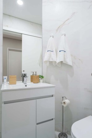 Ein modernes Badezimmer mit einem weißen Waschtisch, einem Spiegel, zwei an der Wand hängenden Handtüchern, einem Toilettenpapierhalter und einer kleinen Topfpflanze.