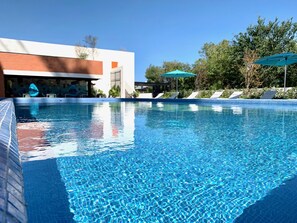 A clear swimming pool with lounge chairs and umbrellas, surrounded by greenery and a modern building.