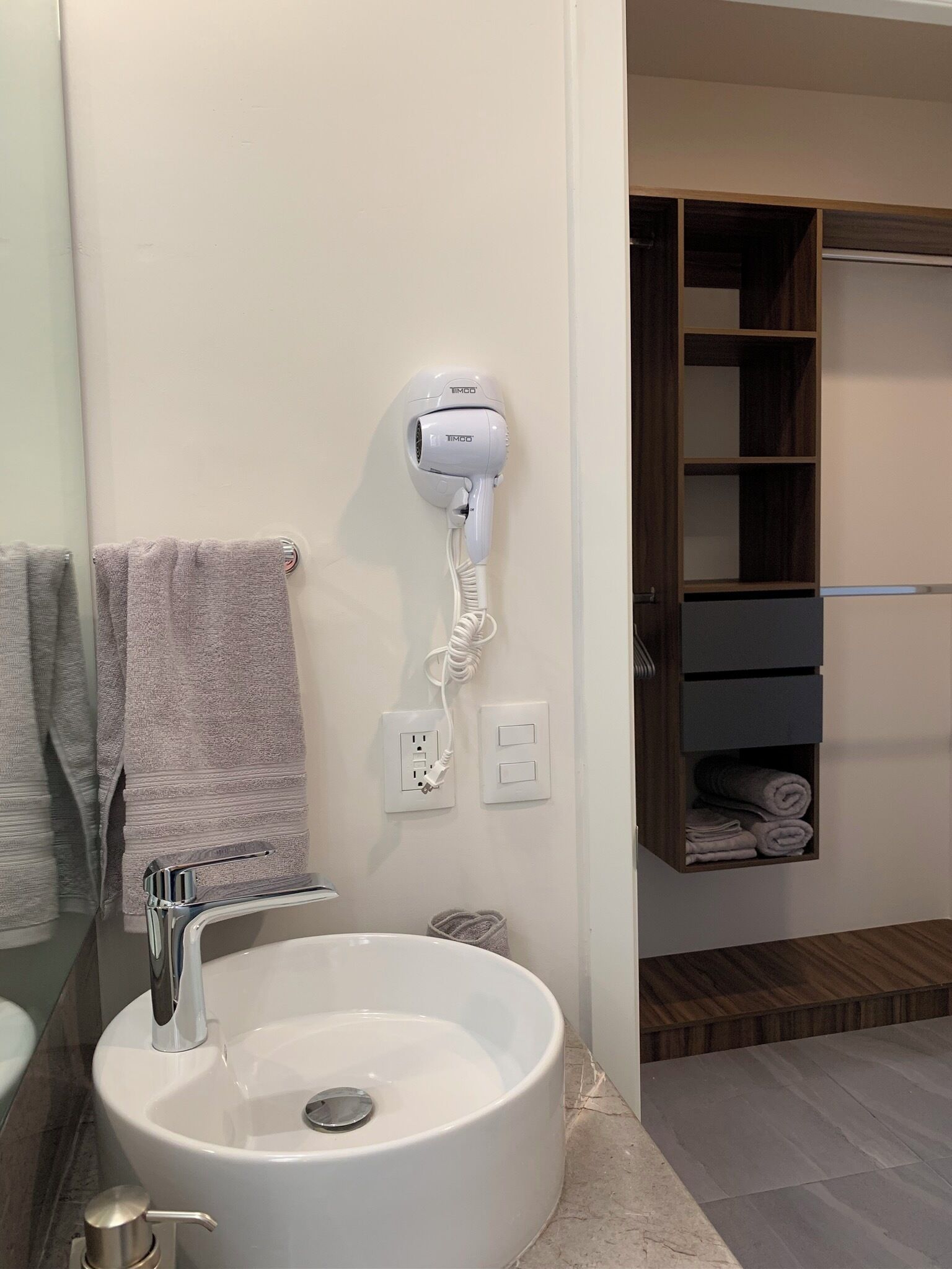A bathroom with a white sink, a towel rack, a hairdryer mounted on the wall, and a wooden shelf with folded towels.