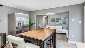 A modern kitchen with a stainless steel refrigerator, a wooden dining table, and a white sofa in the adjacent living area.