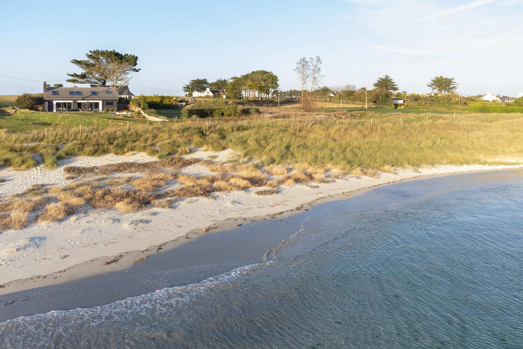 Une propriété située sur la côte, dotée d’une plage de sable, de dunes herbeuses et d’une grande maison avec un toit en pente.