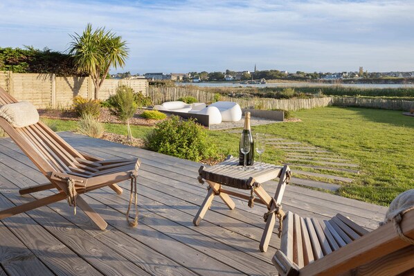 A wooden deck with two lounge chairs and a bottle of champagne, overlooking a grassy area and a distant town.