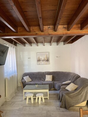 Una sala de estar acogedora con un sofá seccional gris, una mesita blanca con taburetes, vigas de madera en el techo y un cuadro en la pared.
