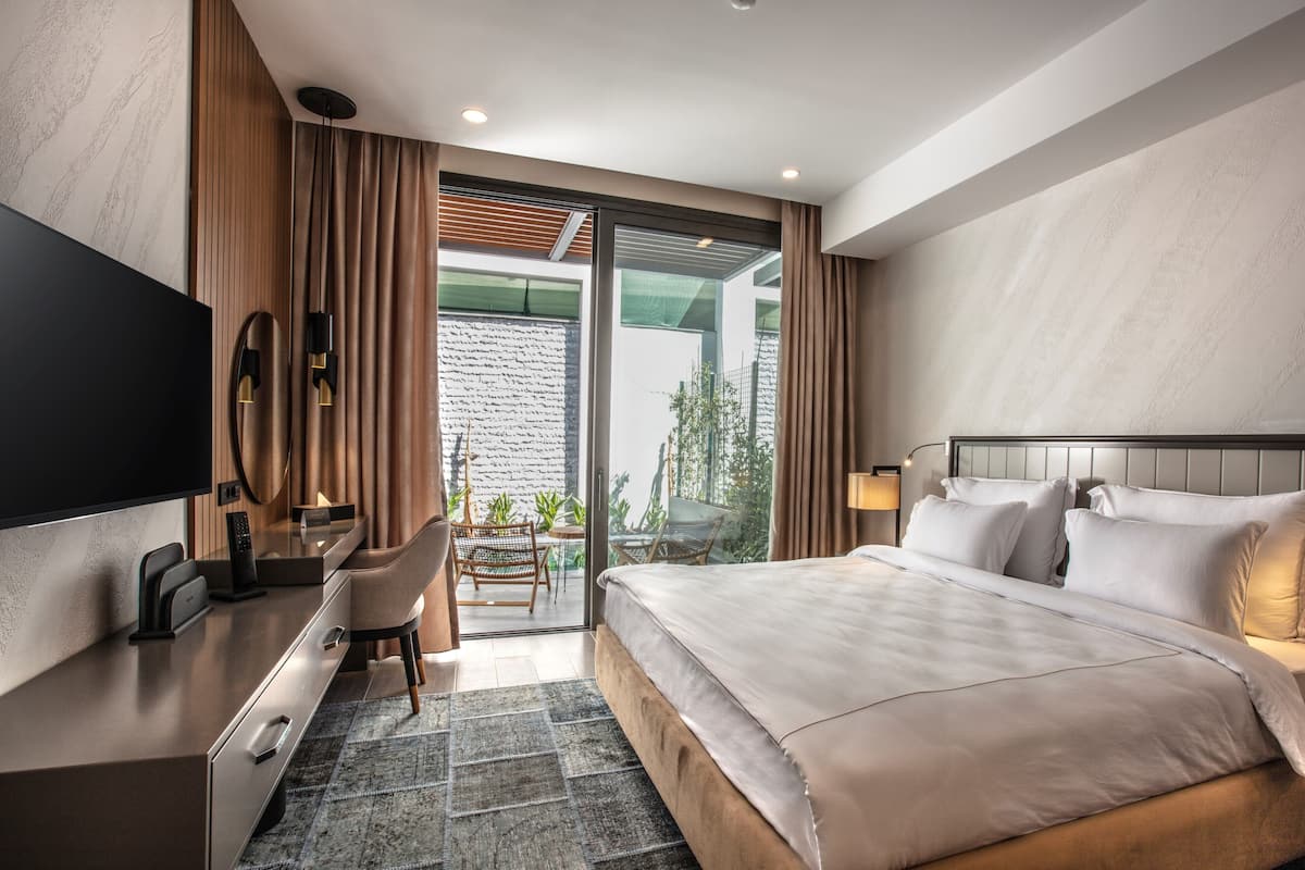 Quarto de hotel moderno com uma cama grande, uma escrivaninha com televisão de tela plana, uma cadeira e vista pela porta de vidro.