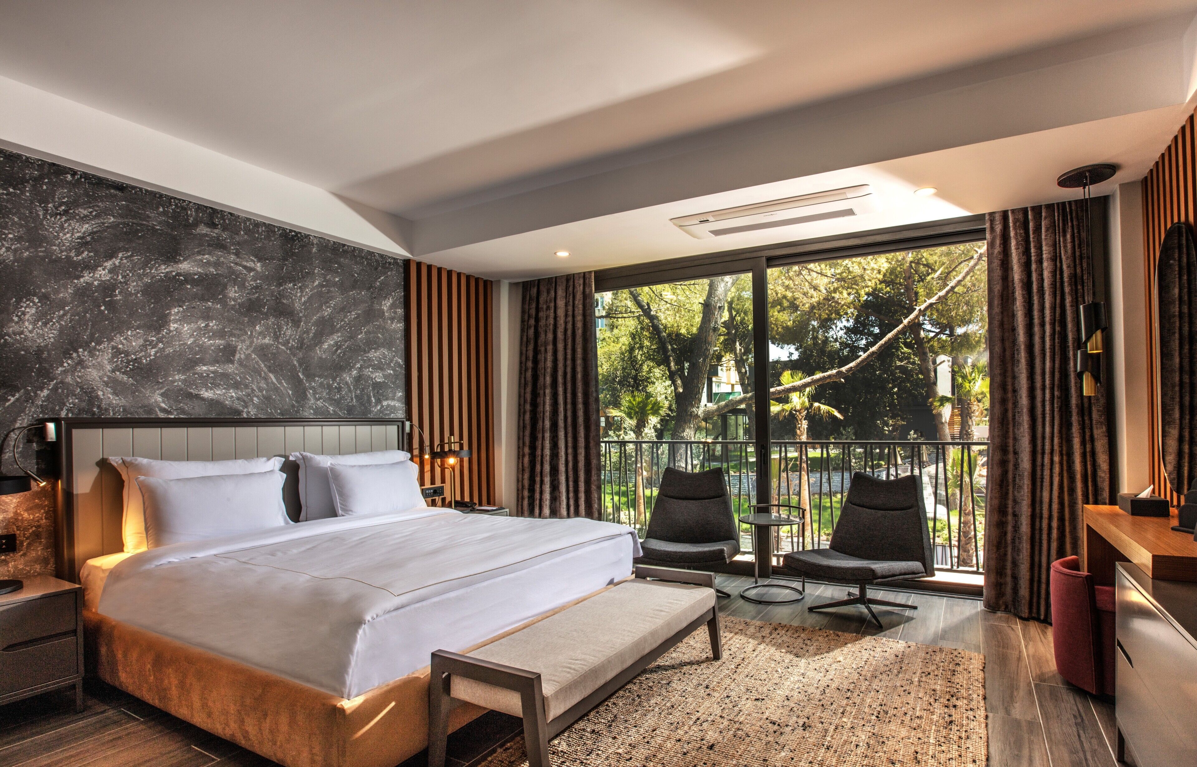 Una habitación de hotel moderna con una cama grande, un balcón con sillas y vistas a árboles.