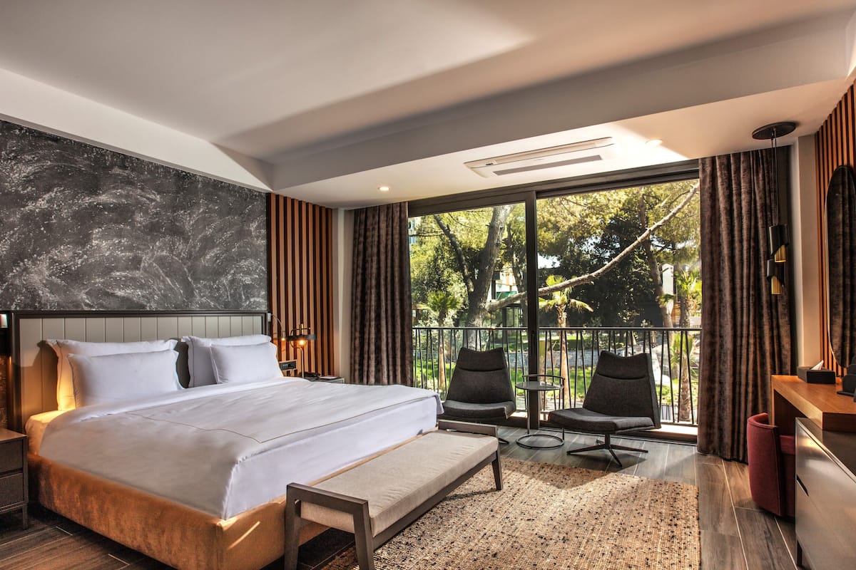 Quarto de hotel moderno com uma cama grande, uma varanda com cadeiras e vista para árvores.
