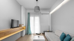 Ein modernes Wohnzimmer mit einem Flachbildfernseher auf einer Holzablage, einem weißen Couchtisch, einer Couch mit türkisfarbenen Kissen und einem großen Fenster mit transparenten Vorhängen.