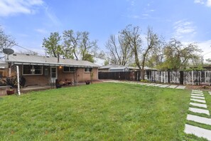 A backyard with a grassy lawn, a brick house, a wooden deck, and a wooden fence. - 5 Bedroom House with Crib - Walk to Downtown Littleton! (Littleton)