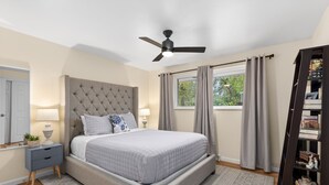 A bedroom with a tufted headboard, a bed with a quilted bedspread, a bedside table with a lamp, a nightstand, a ceiling fan, a window with curtains, and a bookshelf.