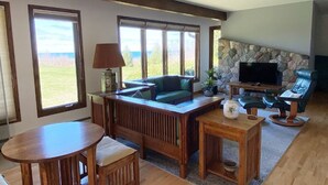 Ein gemütliches Wohnzimmer mit Kamin aus Stein, einem Fernseher und Blick ins Freie durch große Fenster.
